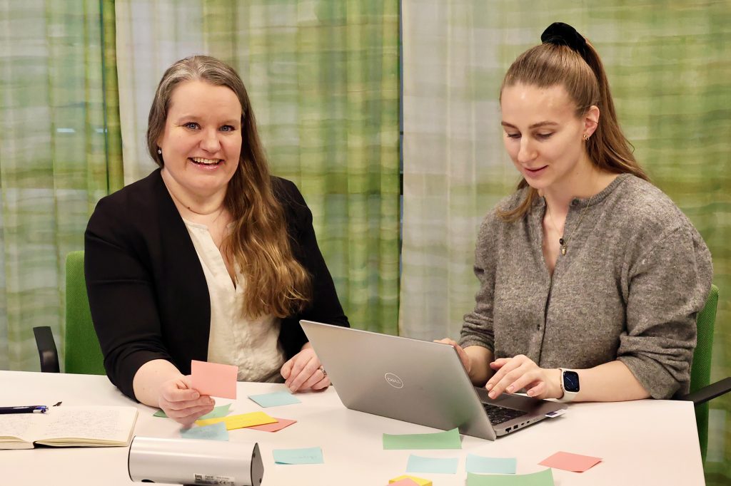 Elsi Vuohelainen katsoo kameraan hymyillen. Hänen kollegansa Eevi-Marie Tihinen kirjoittaa muistiinpanoja tietokoneella.