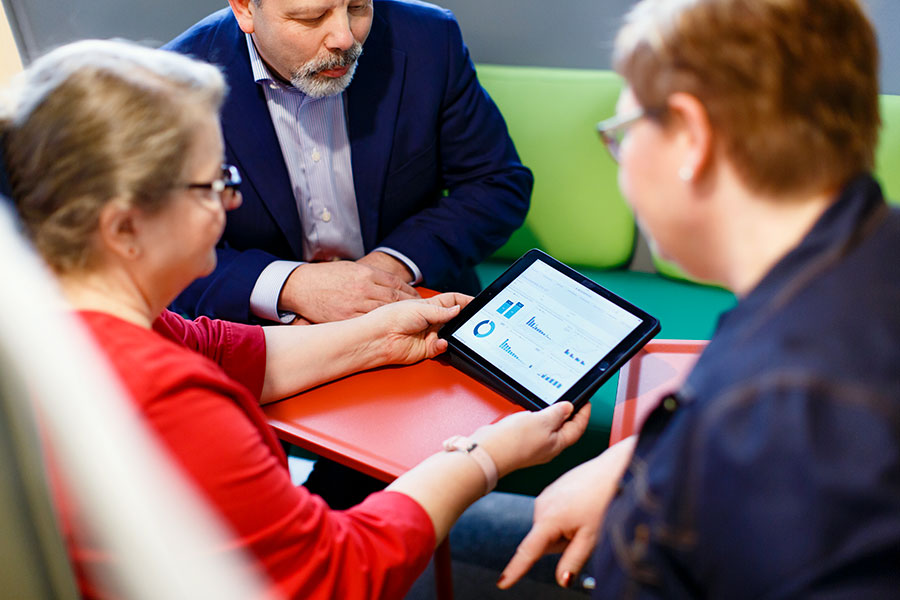 Keski-ikäinen mies ja kaksi naista tutkivat yhdessä tabletilta tilastotietoja.